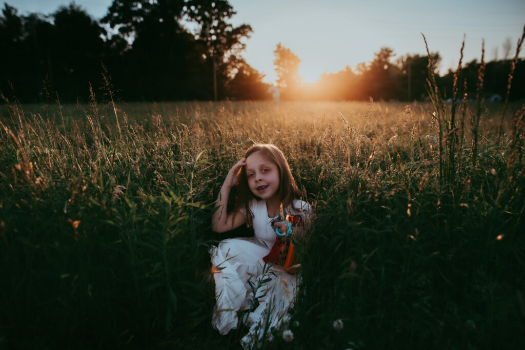 little girl sitting in tall grass smiling with sunset behind her