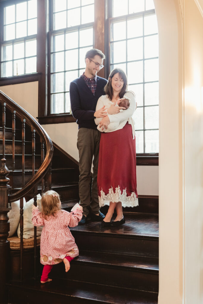 mom and dad hold newborn as toddler attempts to climb stairs nearby