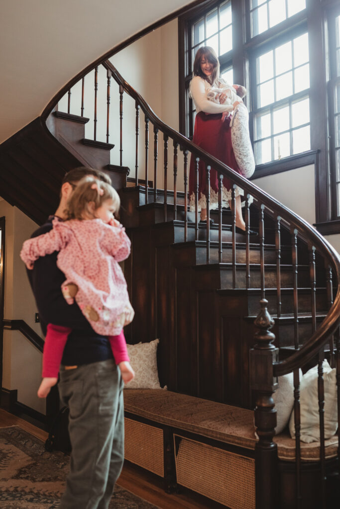 mom descends the staircase with newborn while dad and toddler watch from below