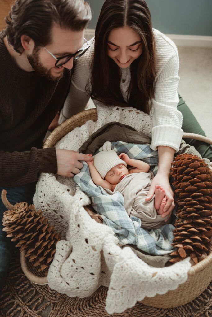 mom and dad holding babys hand and foot as they look down at him in the Moses basket.