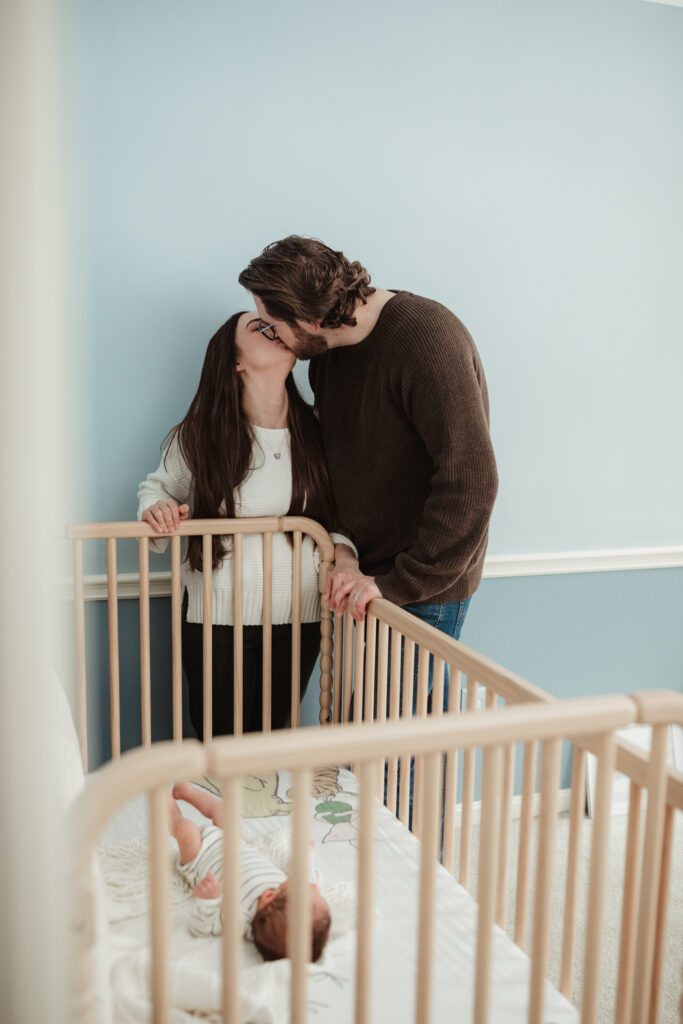 mom and dad kiss while they stand against the baby's crib