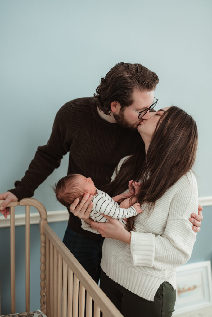 mom and dad kiss as mom holds their baby.