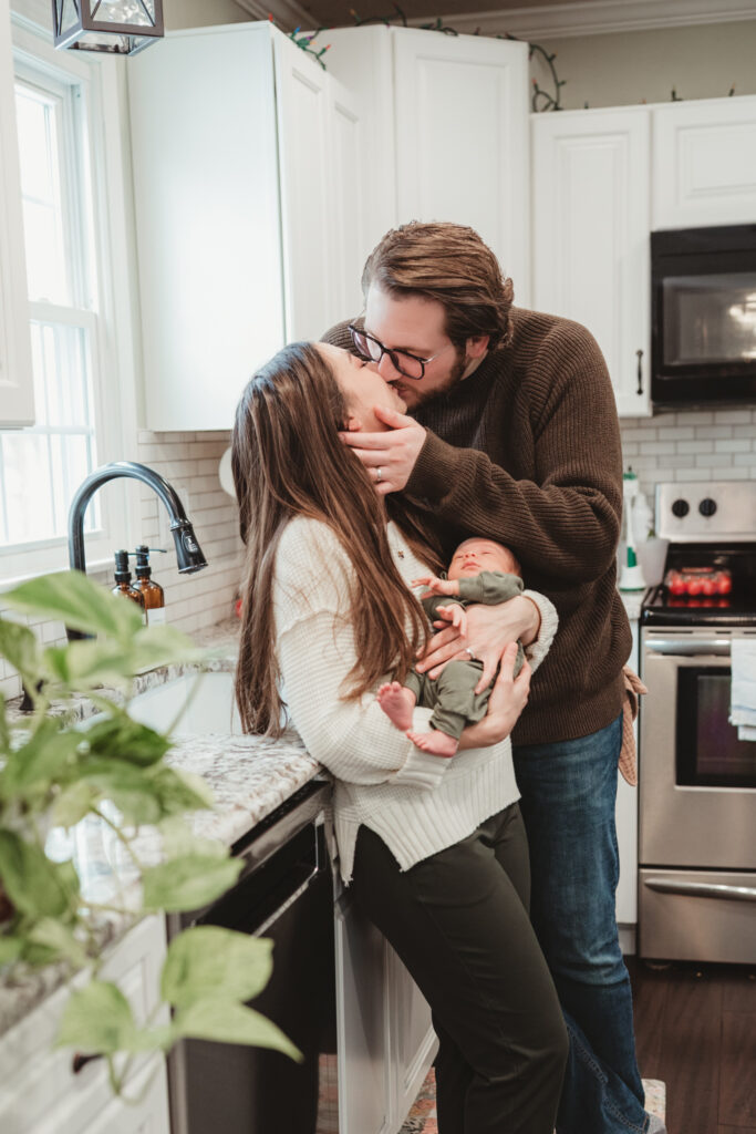 dad kisses mom, who is holding baby, in their kitchen