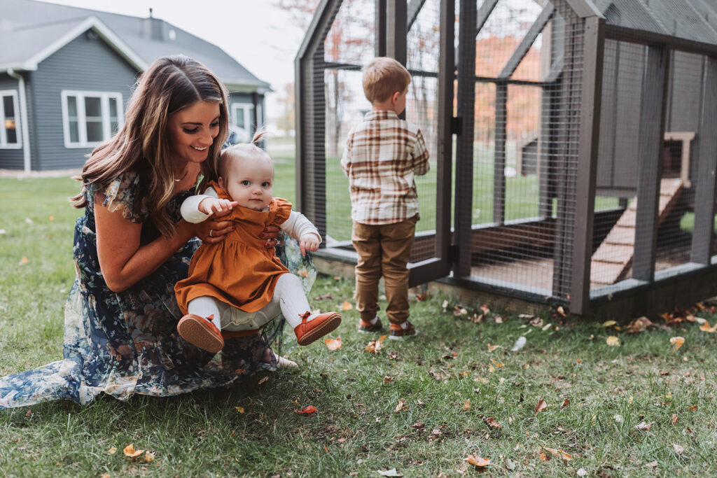mom holding little girl toddler while she kicks her legs, chicken coop and older brother in the background
